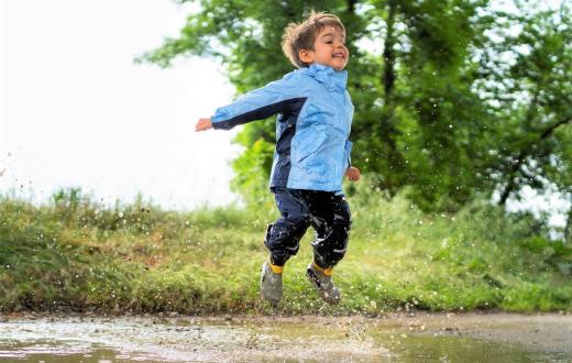 A young boy in a light blue rain jacket, pants and boots jumps gleefully in a mud puddle with green trees and shrubs behind him