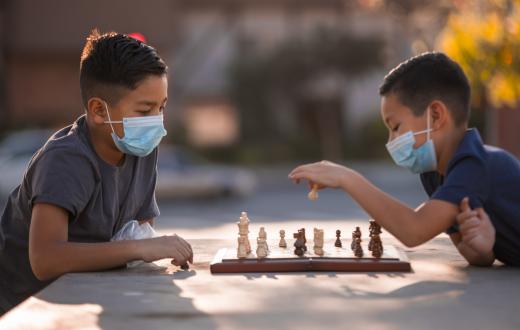 Two boys about age 10 or 11, wearing masks, sitting at a table outdoors playing chess in fall or winter