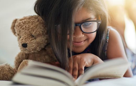 Girl cuddling stuffed bear, smiling and reading a book.