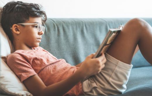 A boy of about 10 wearing glasses sits on the couch and reads a book