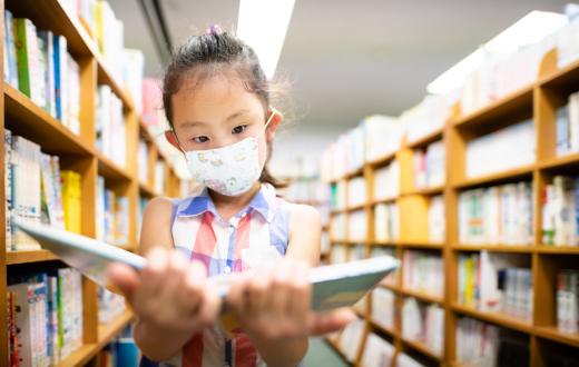 Young Asian girl wearing a mask reads a book in the stacks at a public library
