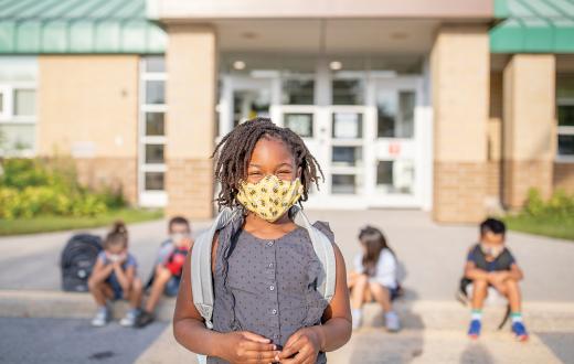boy wearing a mask outdoors with other kids and a school in the background