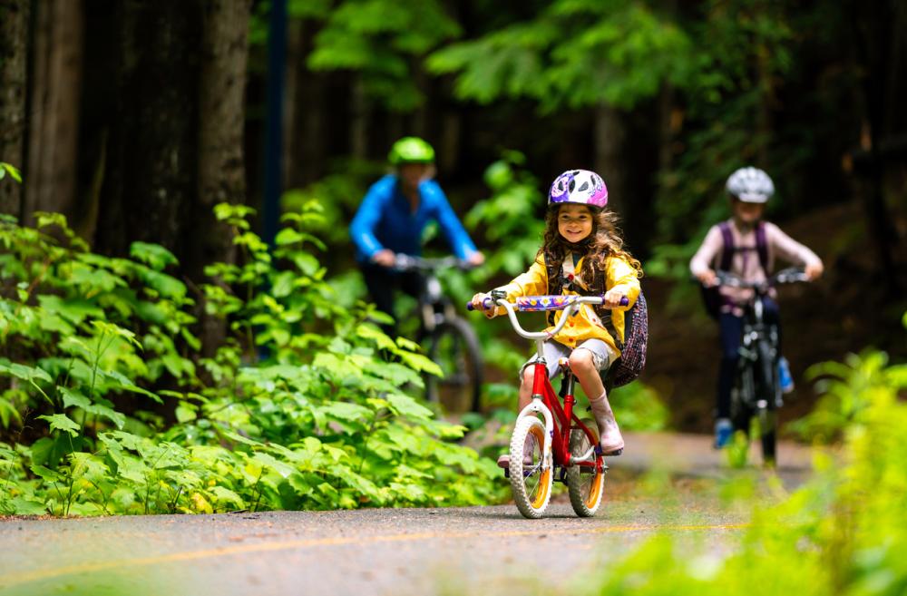 A young girl rides a bike on a bike path with her older sibling and parent following behind. They are wearing helmets and the paved path is lined with green plants indicating summer.