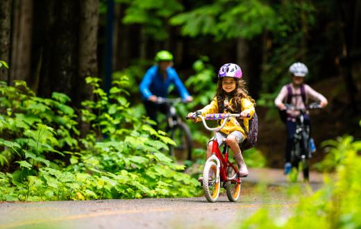 A young girl rides a bike on a bike path with her older sibling and parent following behind. They are wearing helmets and the paved path is lined with green plants indicating summer.